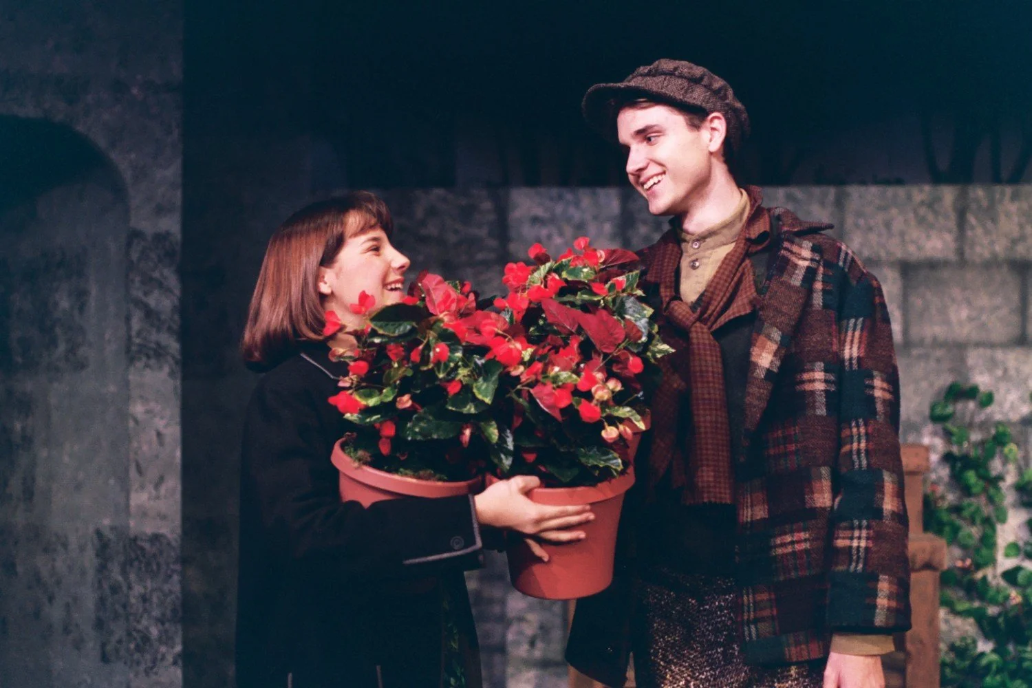 Two people holding pots of red flowers, smiling at each other, dressed in warm, casual clothing.