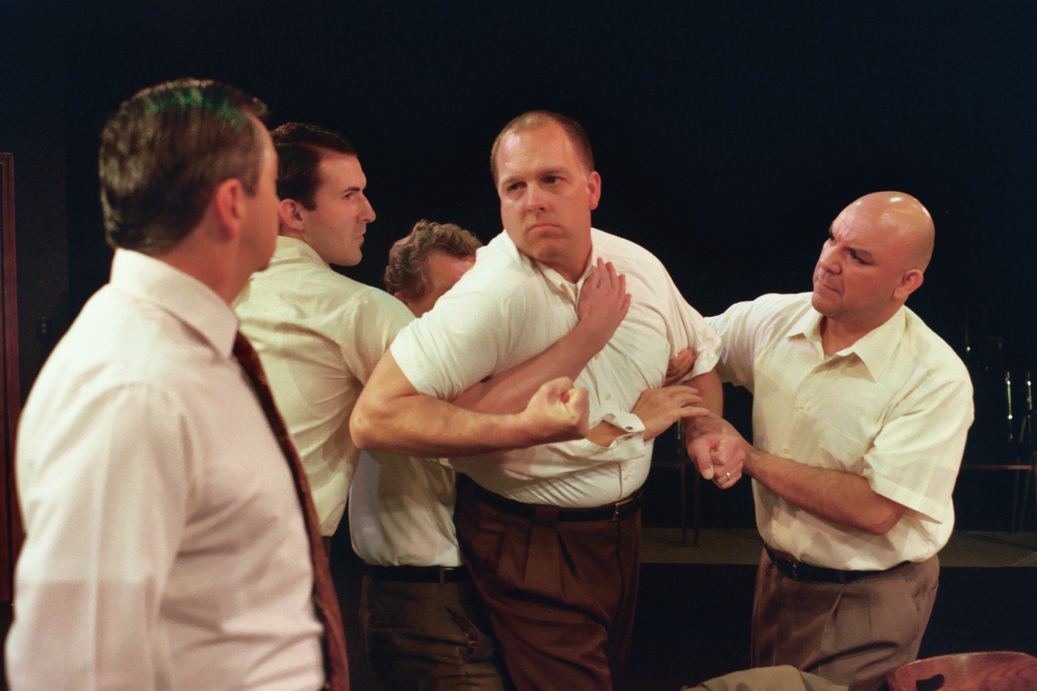 Stage scene with five men in white shirts engaged in a tense confrontation.