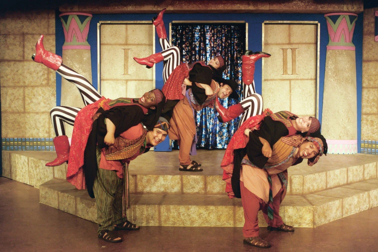 Dancers posing on stage, wearing colorful costumes and headdresses, with striped leggings and red boots, against a theater set backdrop.