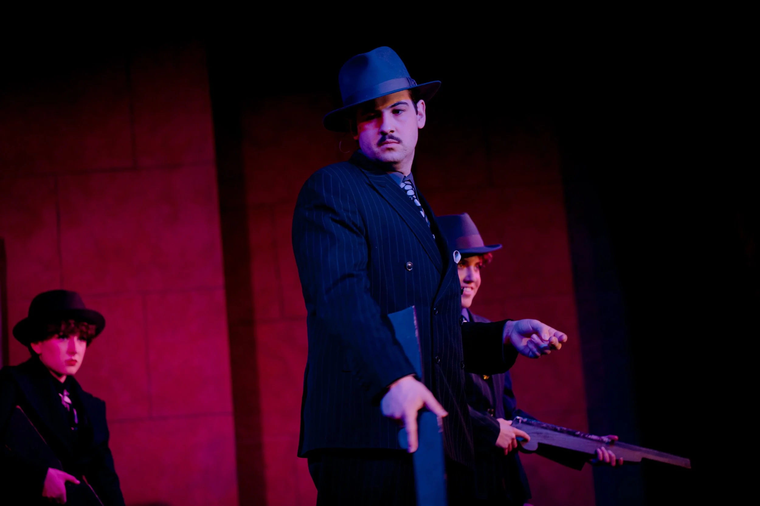 Group of actors in vintage gangster costumes, holding prop guns, performing on stage with dramatic lighting.