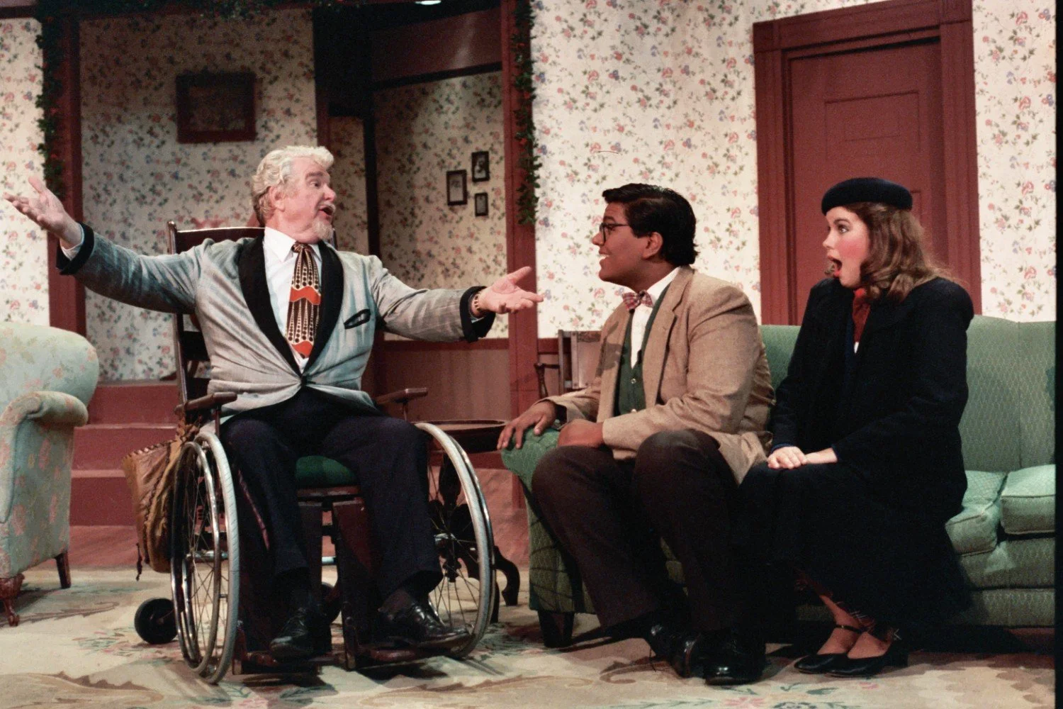 Three actors in a stage play scene, with one man in a wheelchair gesturing dramatically, seated next to a man and a woman on a couch. The background includes floral wallpaper and a closed door.
