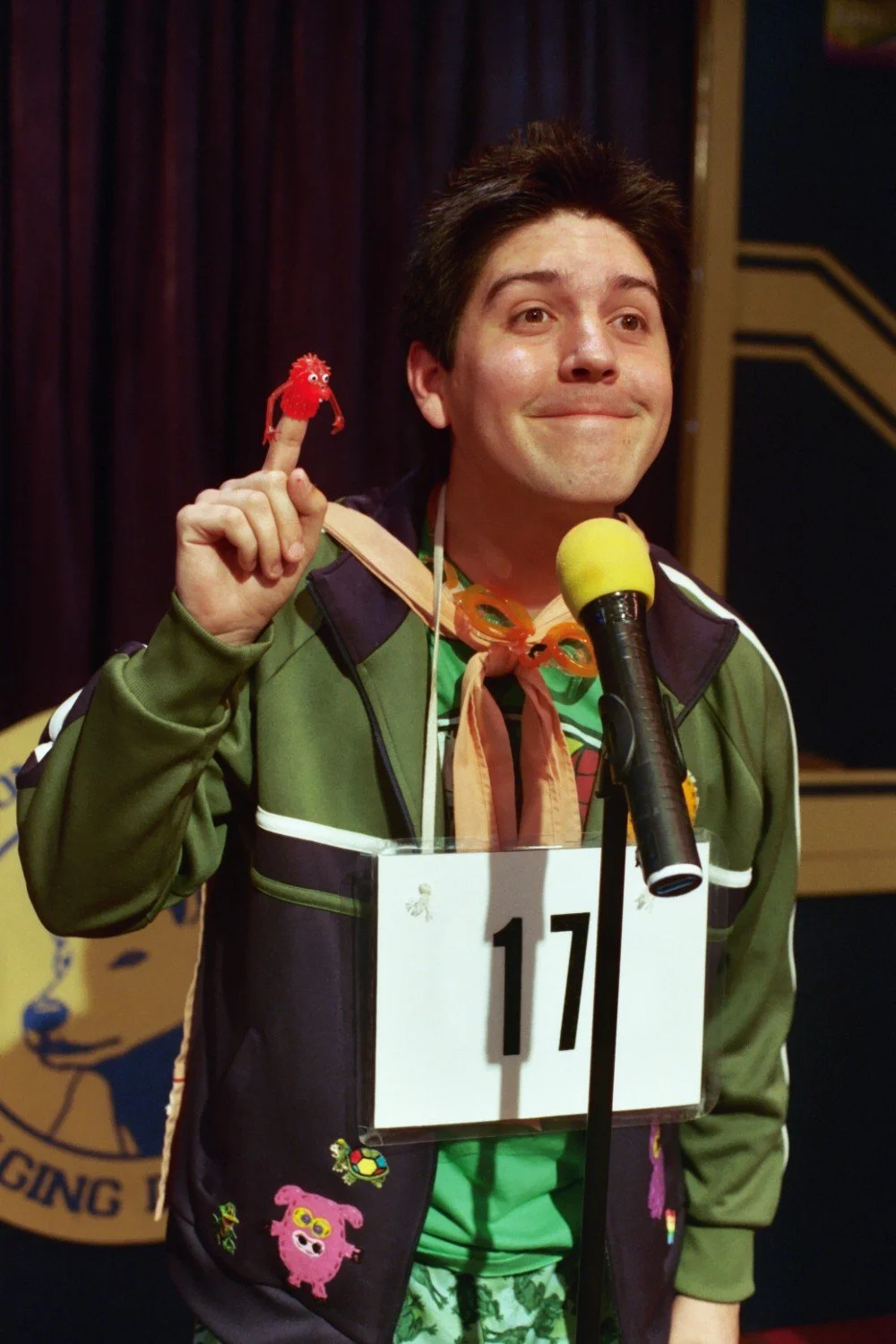 Person at a microphone wearing a costume with the number 17, holding a small red toy creature, and smiling.