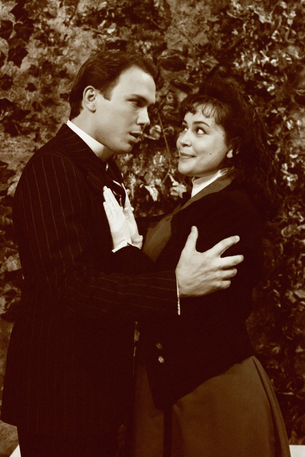 A vintage-style photograph of a man and woman in elegant attire, embracing and gazing at each other in a romantic setting with foliage in the background. The image has a sepia tone.
