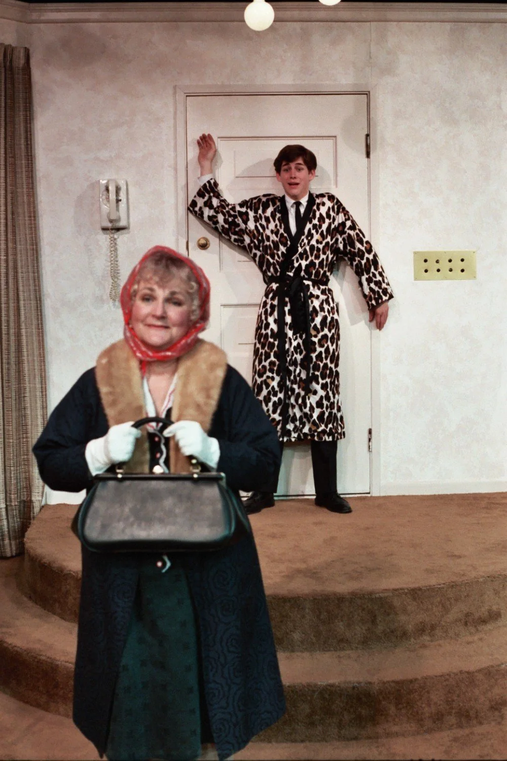 An older woman in a headscarf and coat holds a handbag, while a man in a leopard print robe, tie, and socks stands against a door in the background. The setting appears to be a room with a phone on the wall.