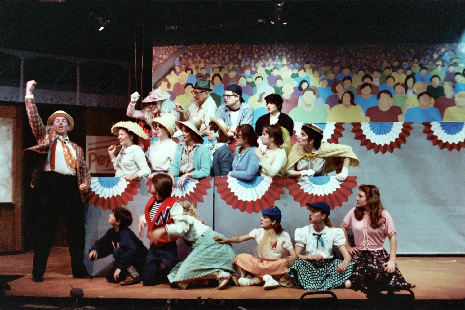 Theatrical performance featuring actors in vintage costumes and hats on a stage with a backdrop of a painted audience, with red, white, and blue decorations.