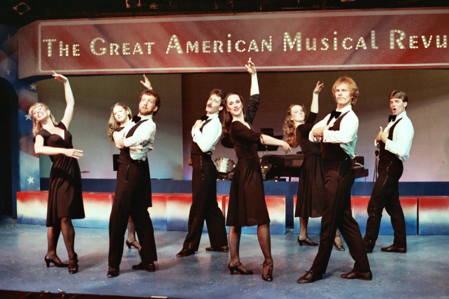 Performers in a musical revue on stage with a sign reading 'The Great American Musical Revue,' dressed in formal attire, posing in mid-dance.