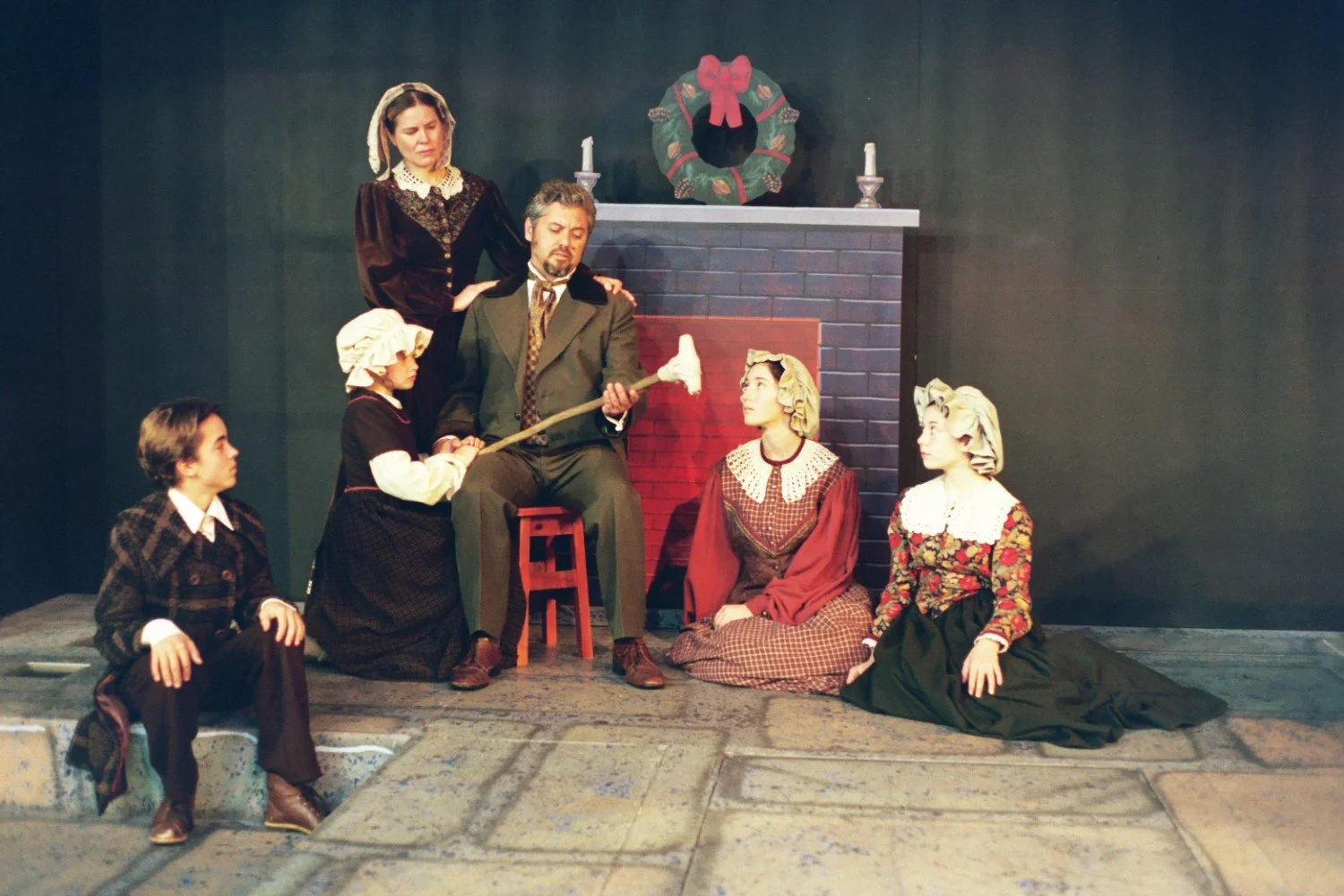 Group of people in Victorian attire sitting by a fake fireplace with a wreath and candles, performing a staged scene.