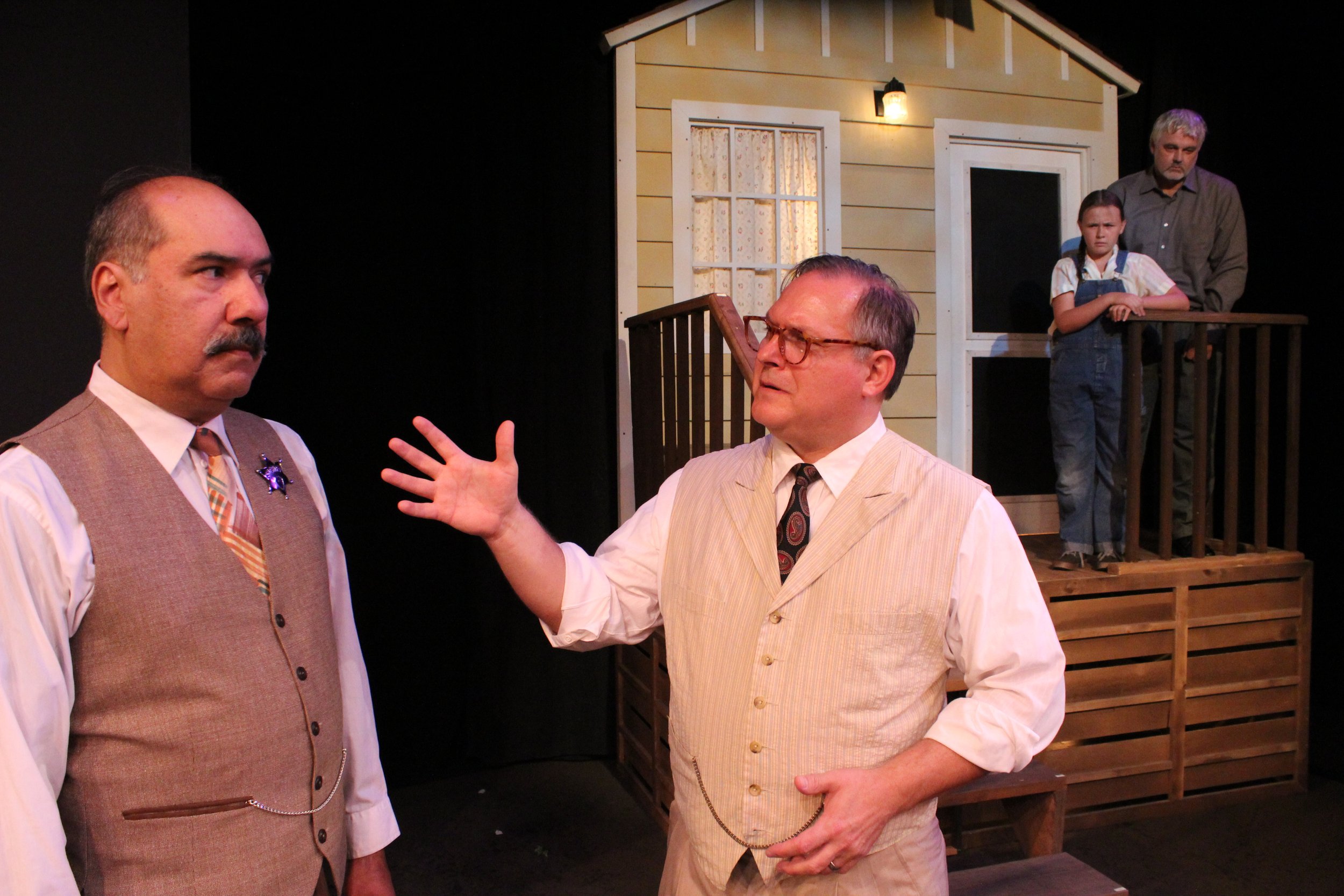 Actors on stage in a dramatic scene with two men in vests having a discussion, while a child and older man watch from a porch of a house set.