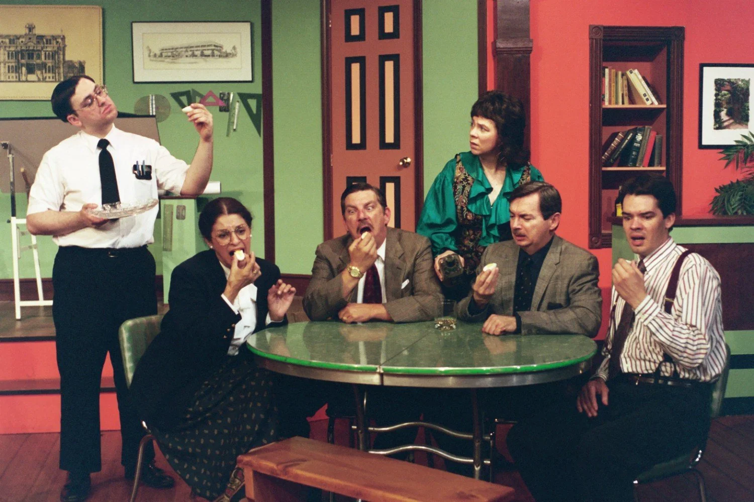 Actors on stage performing a comedic scene, with five people sitting around a table eating, and one standing, holding a tray. The setting appears to be an office or dining room with colorful walls.