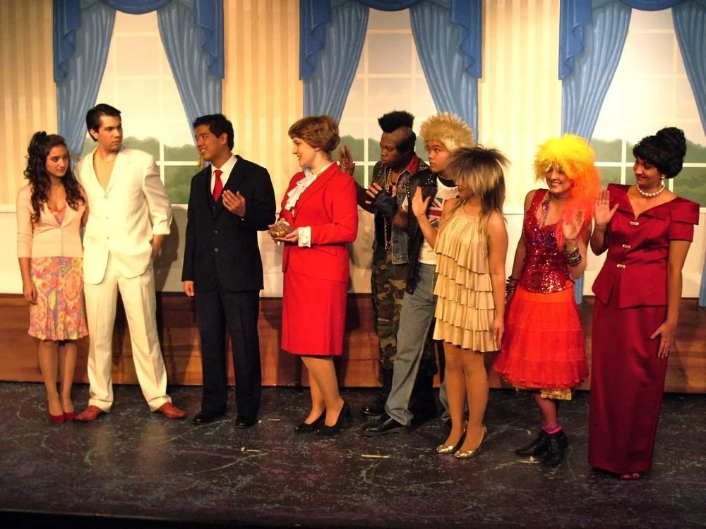 A group of eight people on stage dressed in colorful and varied costumes, standing in front of a theatrical backdrop featuring windows with blue curtains. They appear to be part of a play or theatrical production, engaging in conversation and gesture