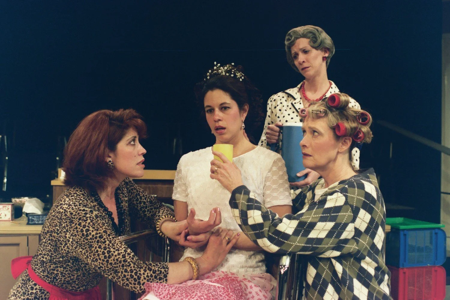 Four women in theater setting, one in a bridal gown, others in casual clothes with hair rollers, engaged in conversation.
