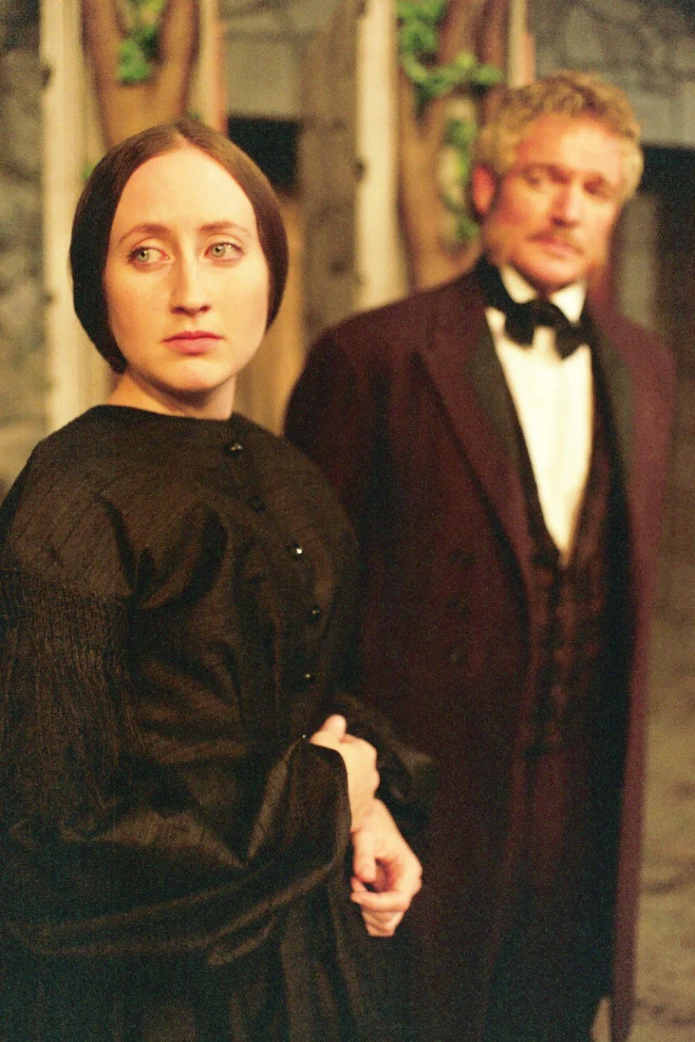 A woman in a dark Victorian-style dress stands in focus, looking to the side, with a man in vintage formal attire blurred in the background. The setting appears to be historical, possibly a play or film scene.