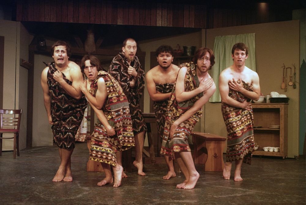 A group of six people on stage performing a comedic scene. They are wearing patterned wrap-around cloth similar to tribal or primitive costumes, standing in humorous poses.