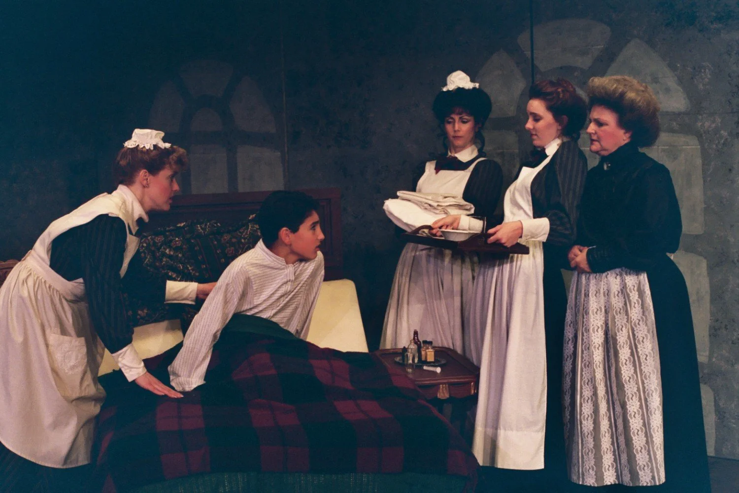 A theatrical scene with a young boy in bed, surrounded by four women in Victorian-style maid uniforms, in a dimly lit room.