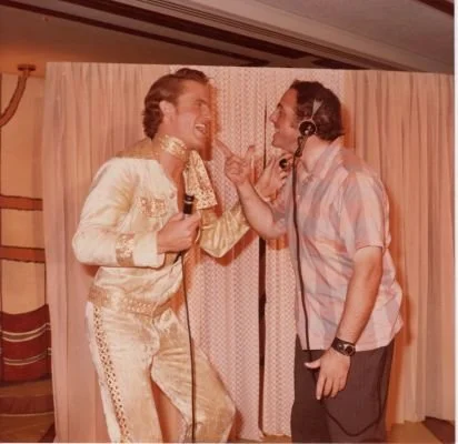 Two men having a fun conversation with microphones, one wearing a gold outfit reminiscent of Elvis Presley, standing in front of a curtain.