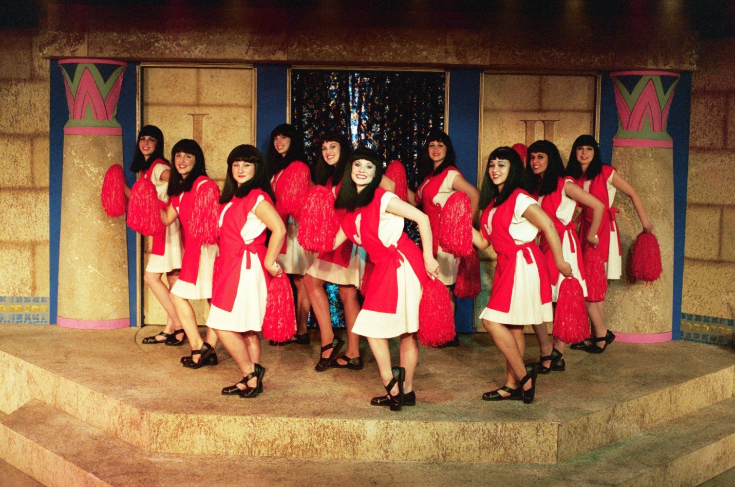 Group of women in matching costumes with red and white dresses and red pom-poms, performing on stage with ancient Egyptian-style decor.
