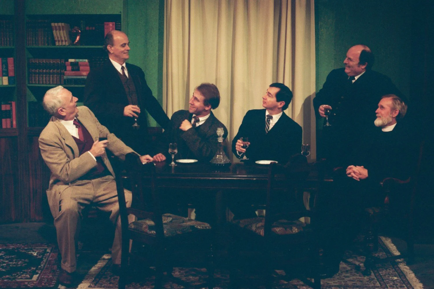 Six men in formal attire sitting and standing around a table in a room with bookshelves and a curtain, holding drinks and in conversation.