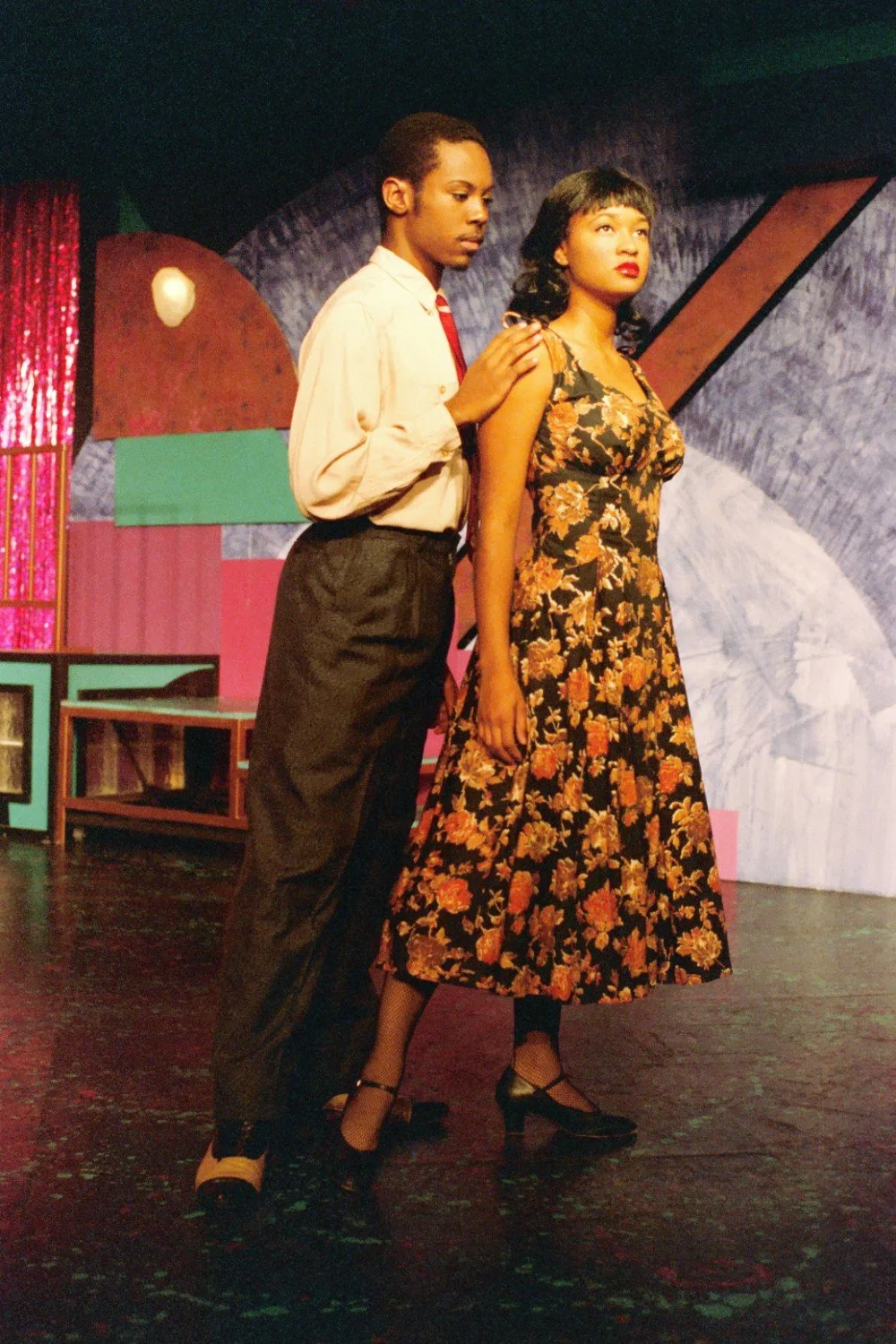 Two performers on a theater stage, one wearing a floral dress and the other in a shirt and tie, with a colorful set in the background.
