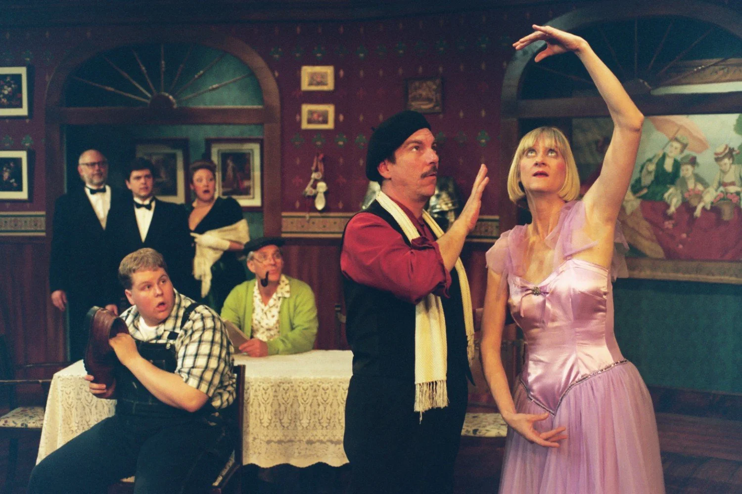 A theatrical scene with seven people in colorful costumes. A man in a beret and scarf gestures dramatically towards a woman in a purple dress who is striking a ballet pose. In the background, five others, including a man in overalls and a person in a