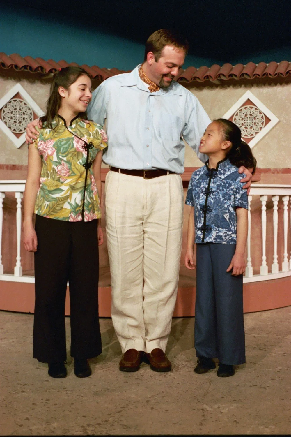 A man with two young girls standing on a stage, smiling and interacting. The man is wearing a light blue shirt and white pants, while the girls are wearing floral patterned shirts and dark pants. The background features a decorative wall with geometr
