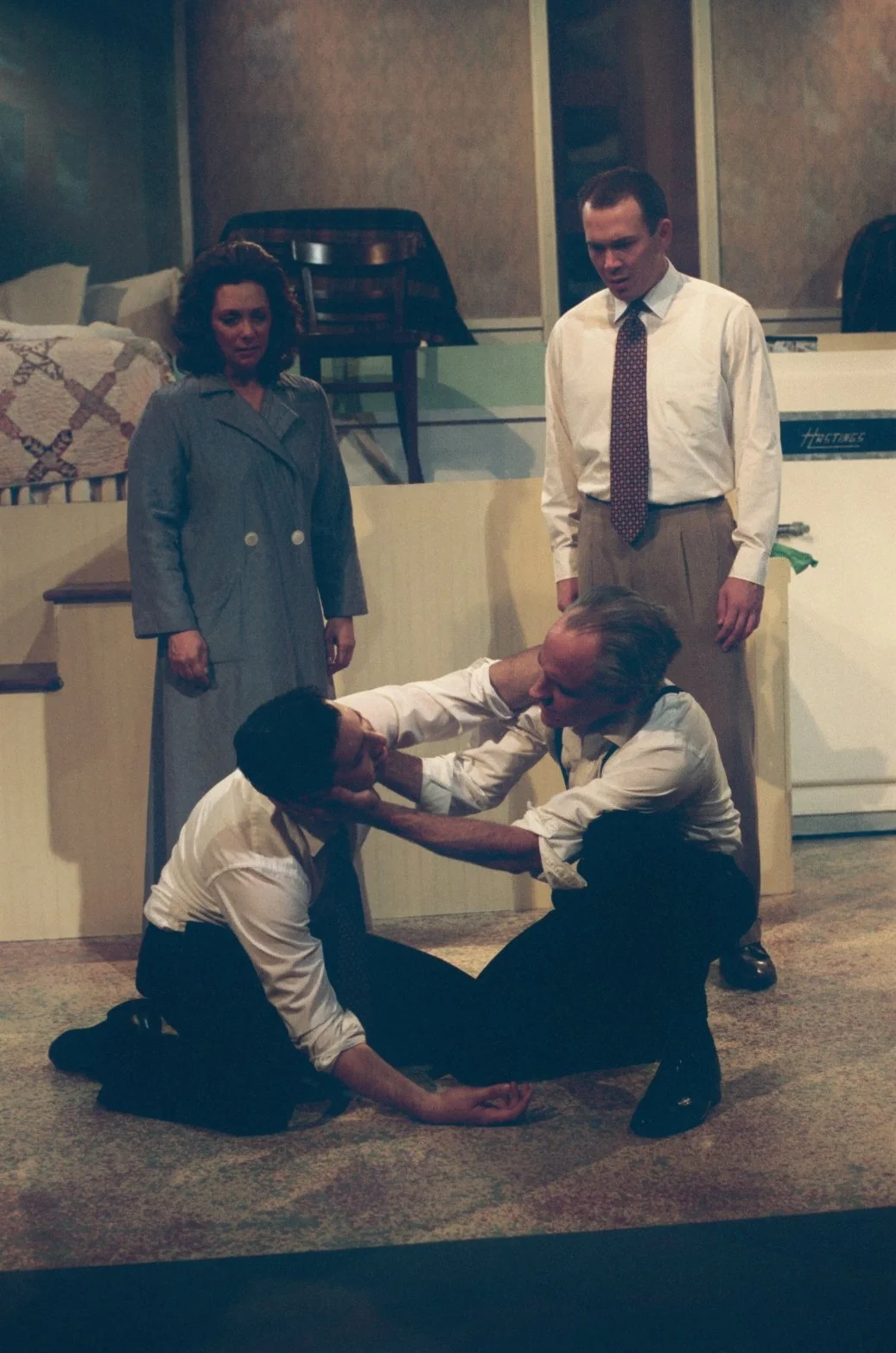Four people in a tense scene, two men on the floor in a struggle, with a woman and another man standing nearby, in a theatrical setting.