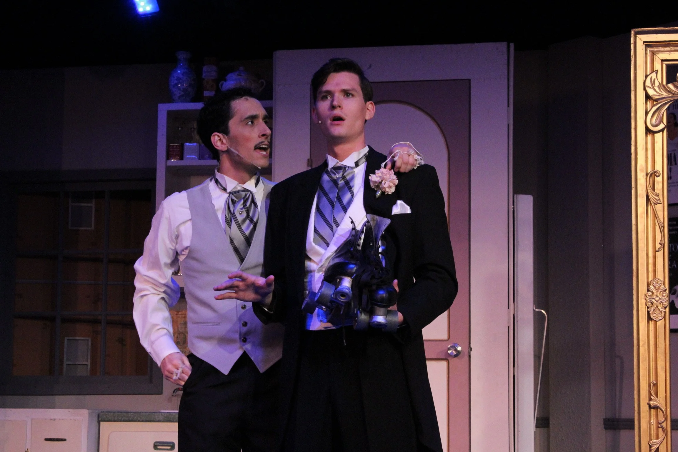 Two men in formal attire; one holding roller skates, staged in a theatrical setting.