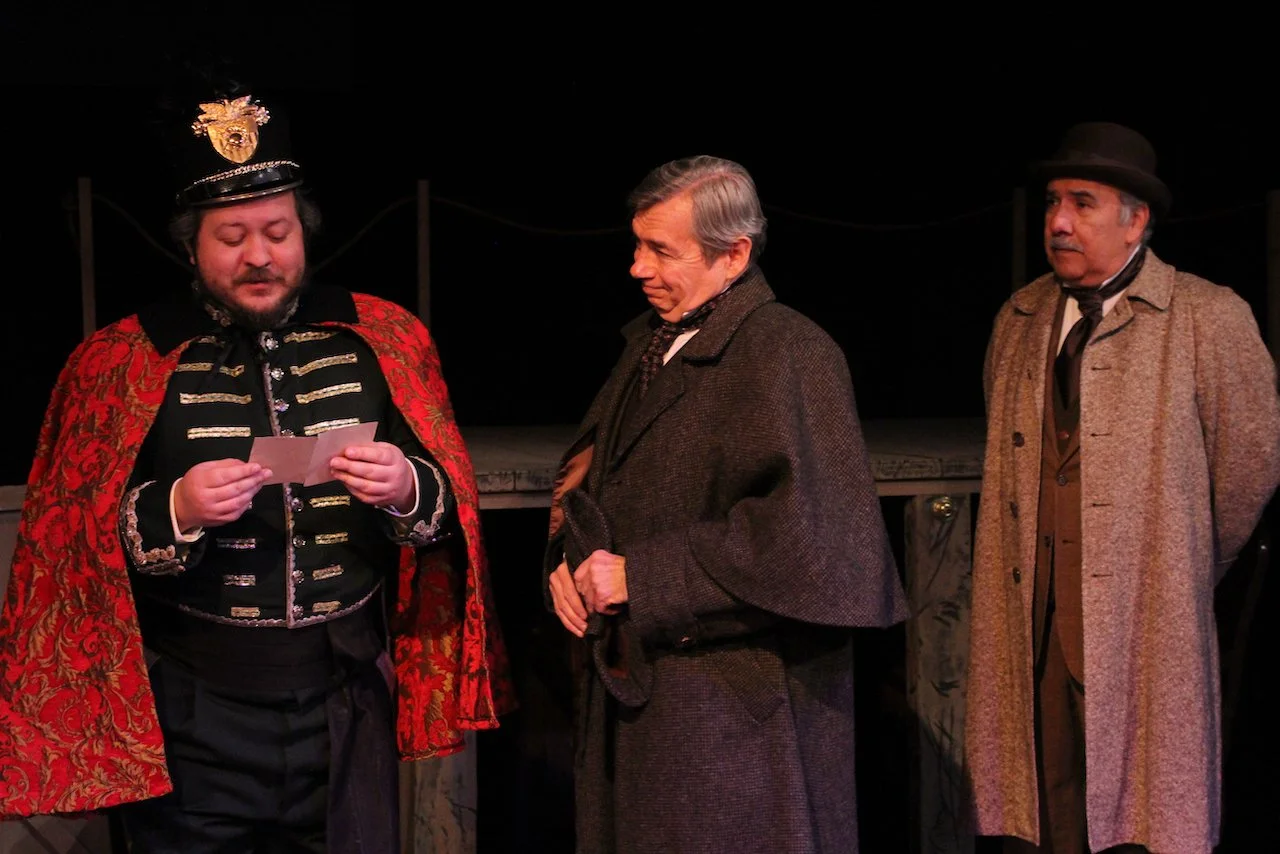 Three men in Victorian-style costumes performing on stage. The man on the left is wearing a red and black ornate military uniform with a cap, holding a piece of paper. The other two men are dressed in period coats and hats, observing.