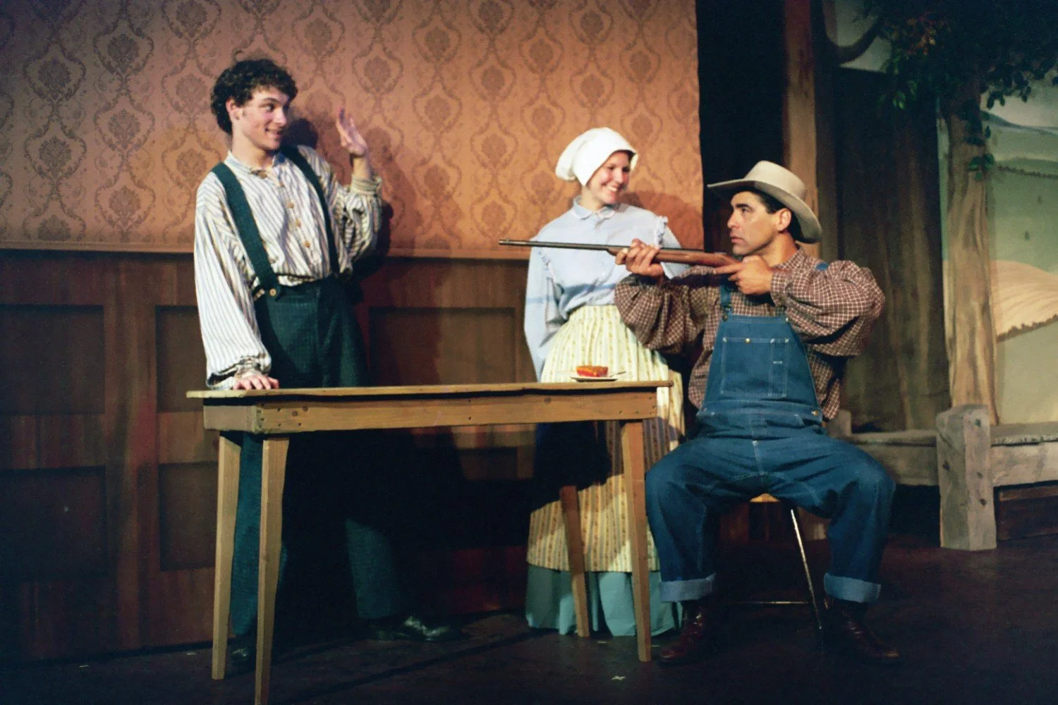 Three people in a theatrical scene wearing 19th-century clothing; two are standing, one is sitting aiming a rifle. The room has vintage wallpaper and a wooden table with a small plate.