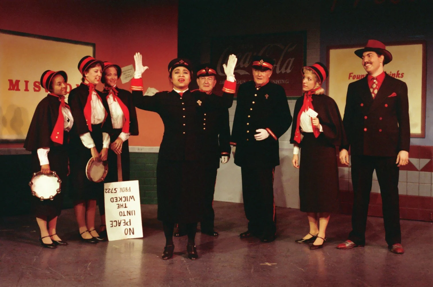 A group of performers in theatrical costumes, including Salvation Army-style uniforms and vintage outfits, standing on stage. One person is holding a tambourine, and a placard with the message 'No Peace Unto The Wicked Prov 57:22' is partially visibl