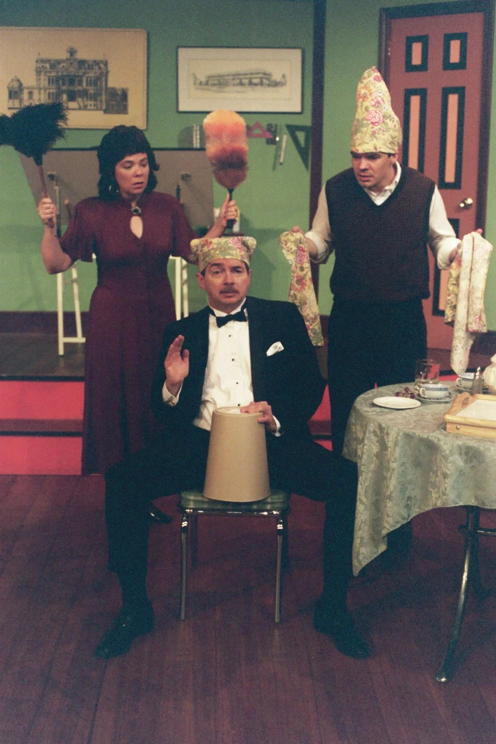 Three people in a comedic scene with whimsical hats, a seated man in formal attire holding a bucket, surrounded by two standing individuals holding dusters, in a room with vintage decor.