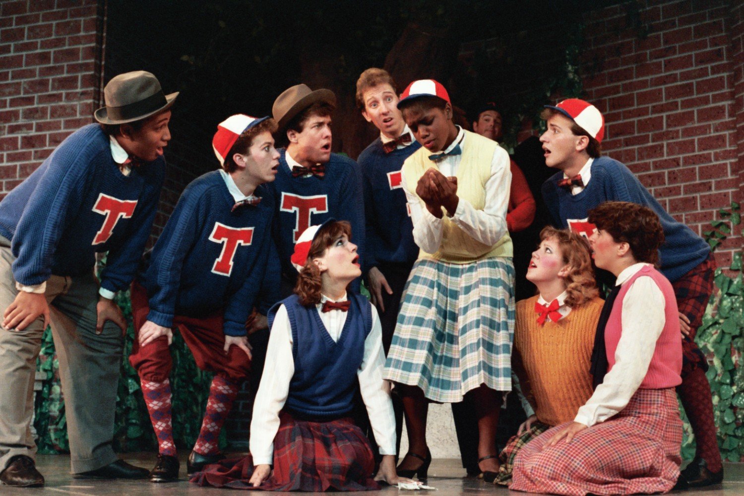 A group of people in vintage-style clothing with letter "T" sweaters and hats, performing on stage with expressions of surprise or intrigue, surrounded by a brick wall and greenery.