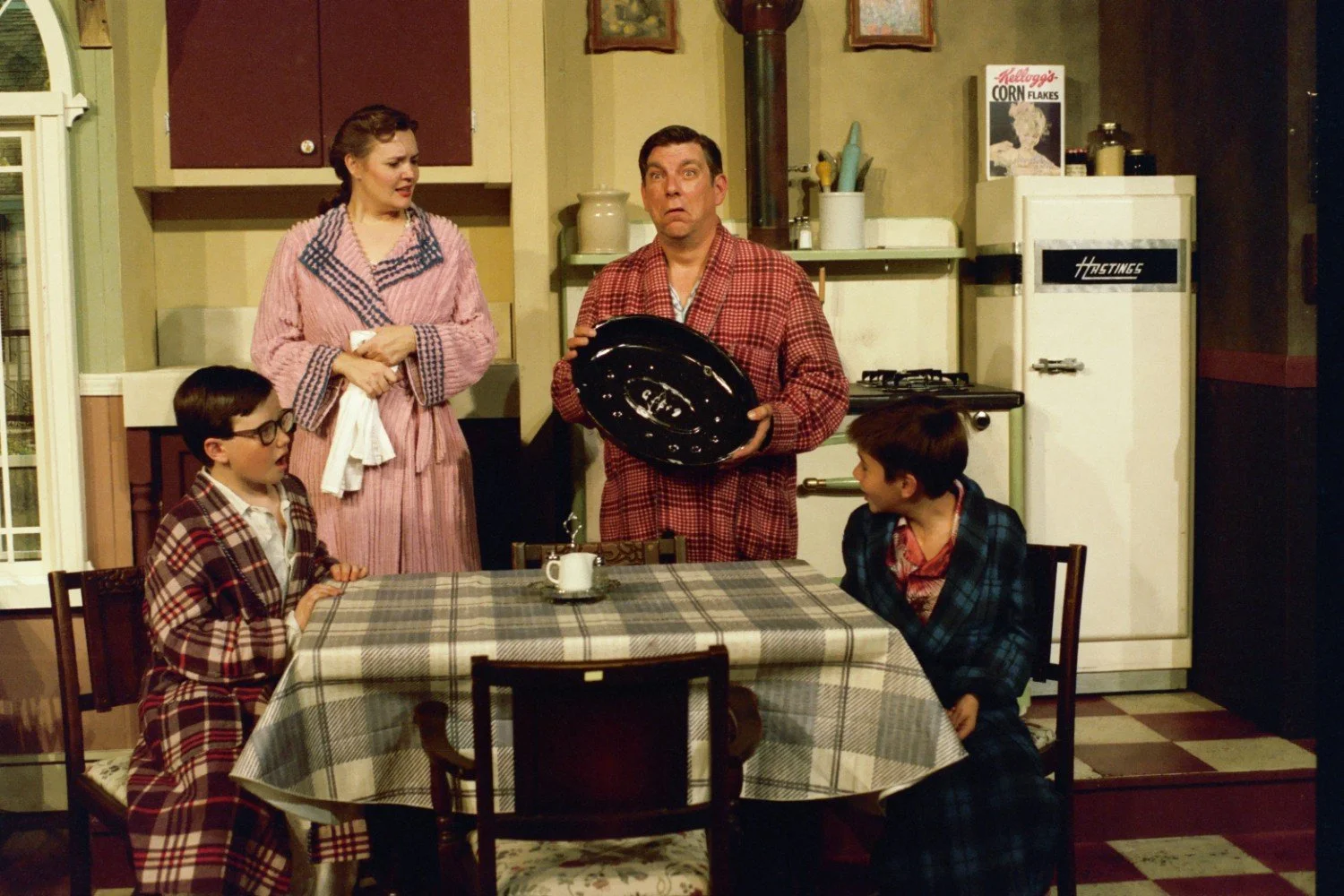 Family in a vintage kitchen, all wearing bathrobes, around a table with a checkered tablecloth. One person holds a large round object, possibly a cookware lid.