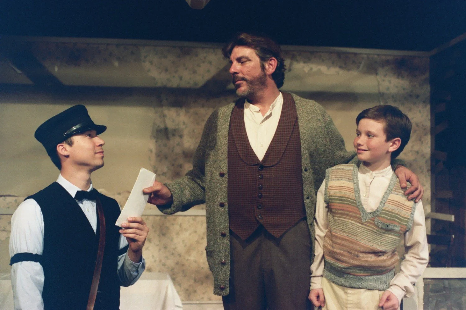 Three actors on stage in a theatrical production. One is dressed in a postman's uniform, handing an envelope to a man in a cardigan and vest. A young boy in a striped vest stands beside them, looking up at the man.