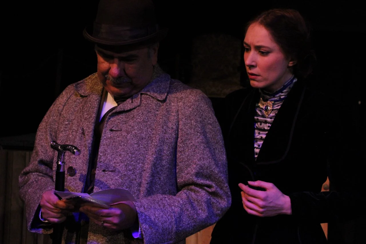 A scene from a theatrical performance with a man in a gray coat and black hat holding papers, standing next to a woman in a dark outfit looking concerned.