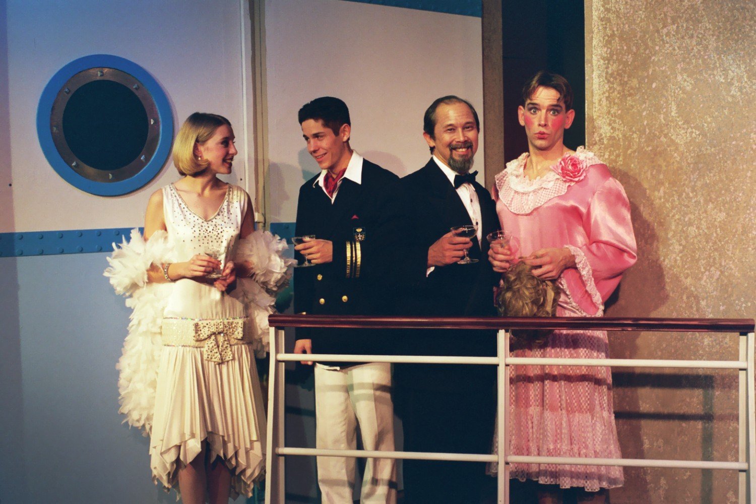 Four people dressed in formal and theatrical attire standing on a ship's deck, holding drinks, with a decorative ship porthole in the background.