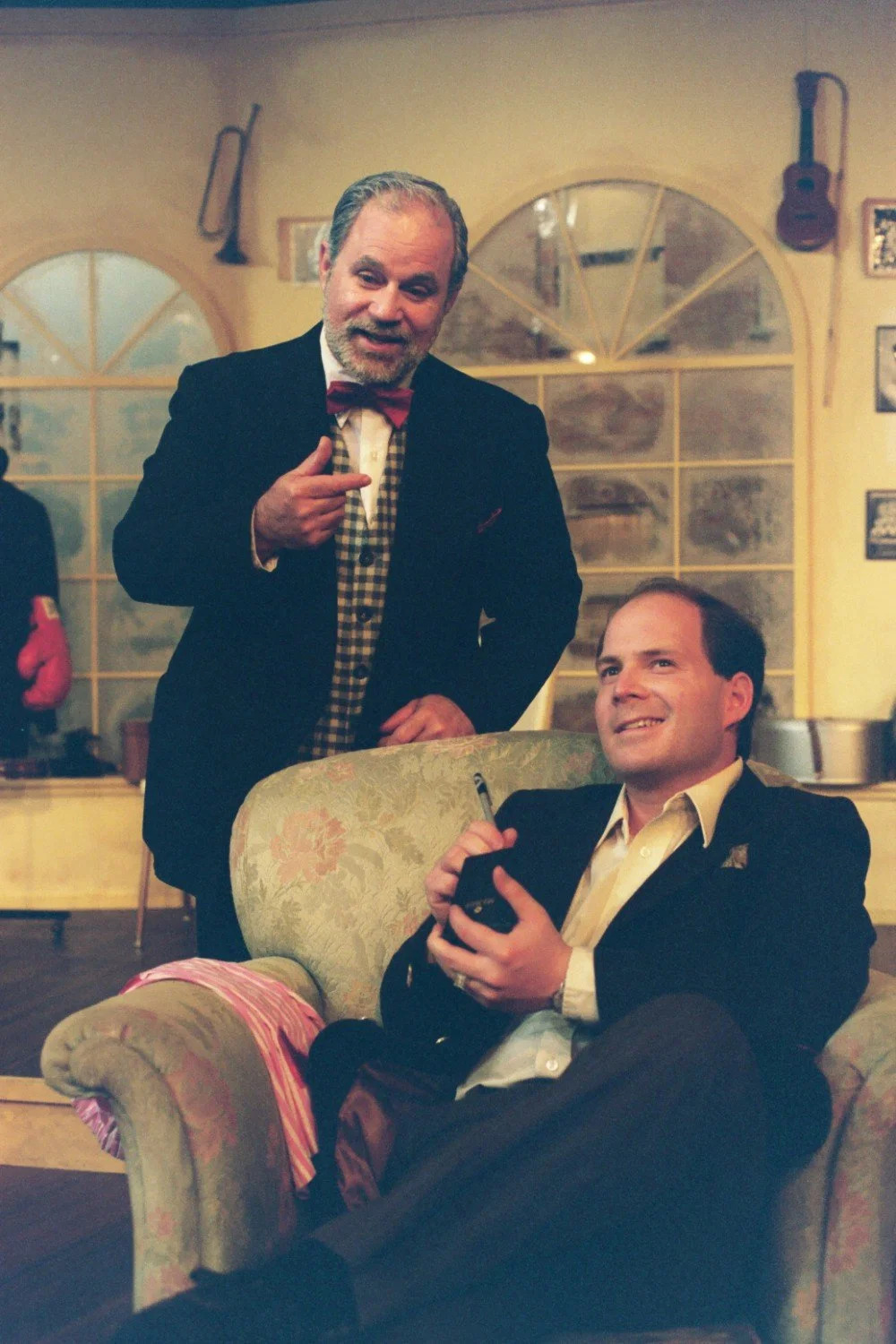 Two men dressed in suits; one standing and gesturing with a pointing hand, and the other sitting on a couch holding a notepad and pen.