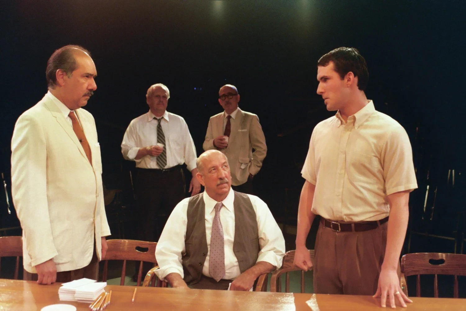 A group of men in formal attire engaged in a serious discussion around a table, possibly during a theatrical performance.