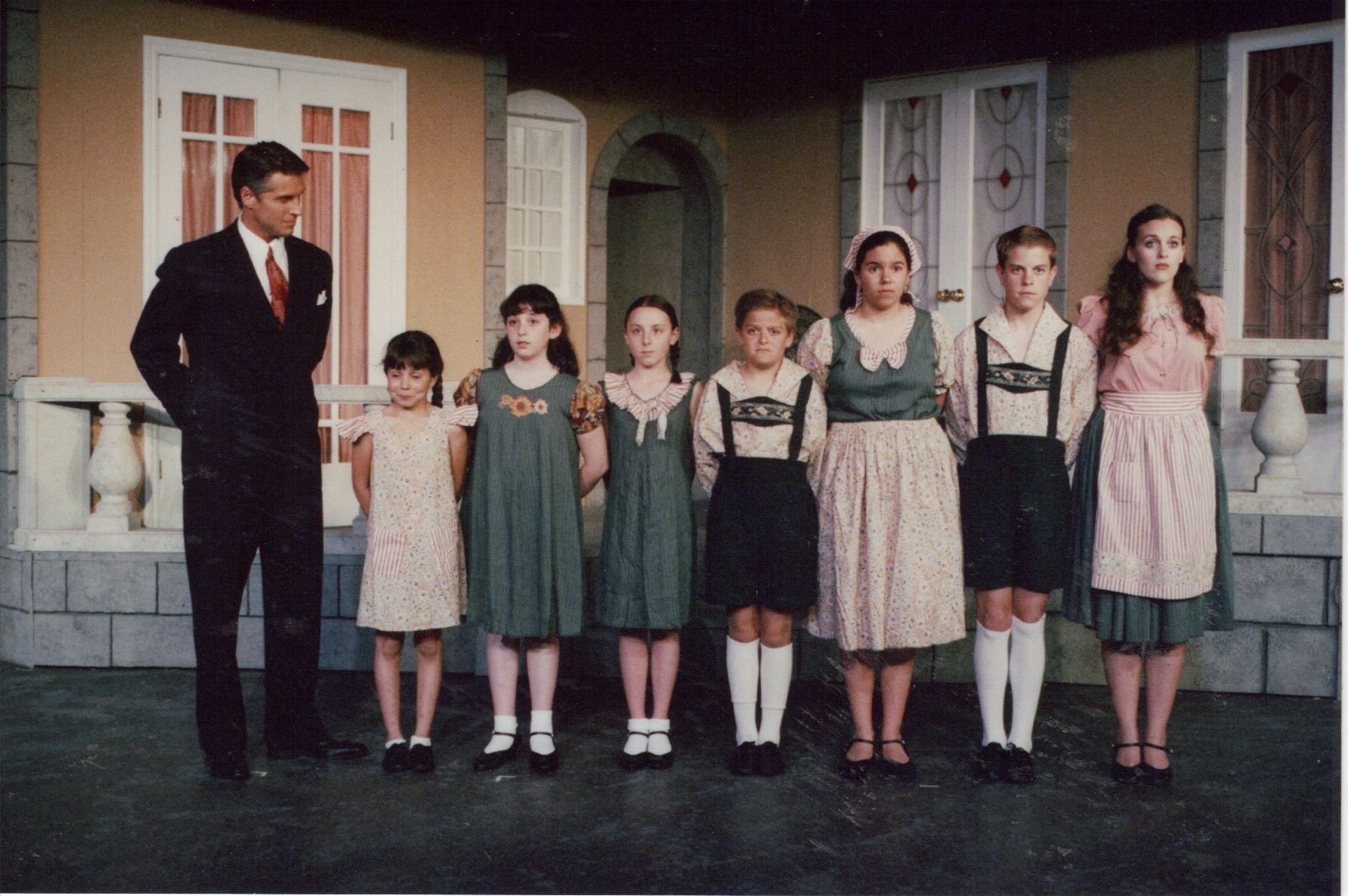 Group of seven children and one adult in period costumes in front of a stage set depicting a house exterior.