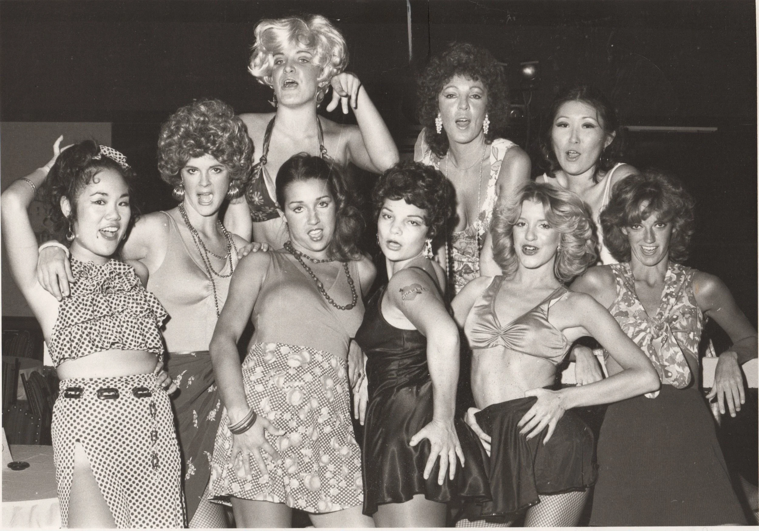 A black and white photo of a group of women posing together. They are wearing vintage-style outfits, including dresses and tops. Some women have their hair styled in curls or waves, and appear to be having fun with expressive poses and facial express