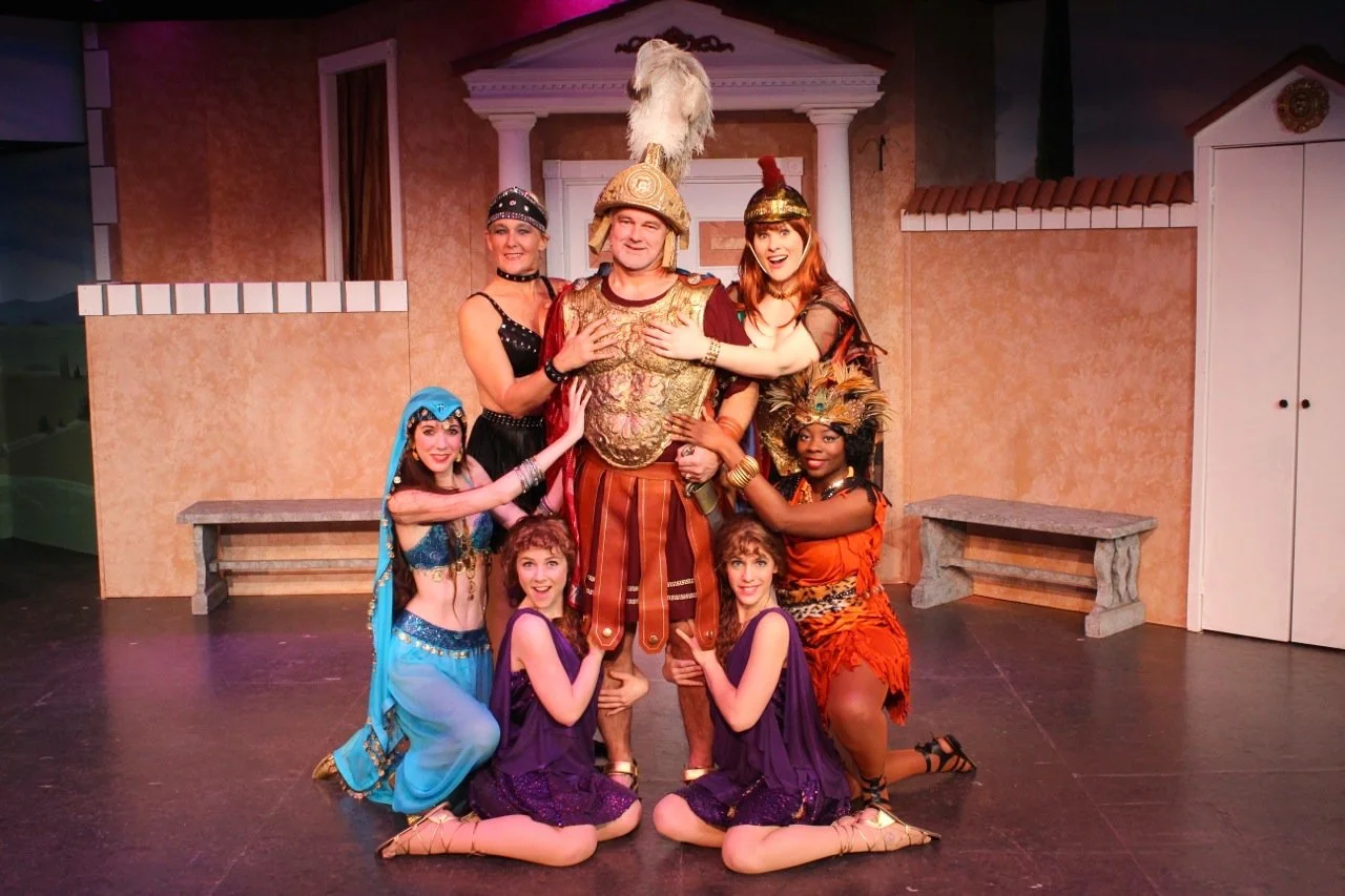 A group of actors in costumes from a theatrical performance resembling ancient Roman or Greek theme. The central figure is dressed as a Roman soldier, surrounded by women in various exotic costumes, including a dancer in blue and others in colorful o
