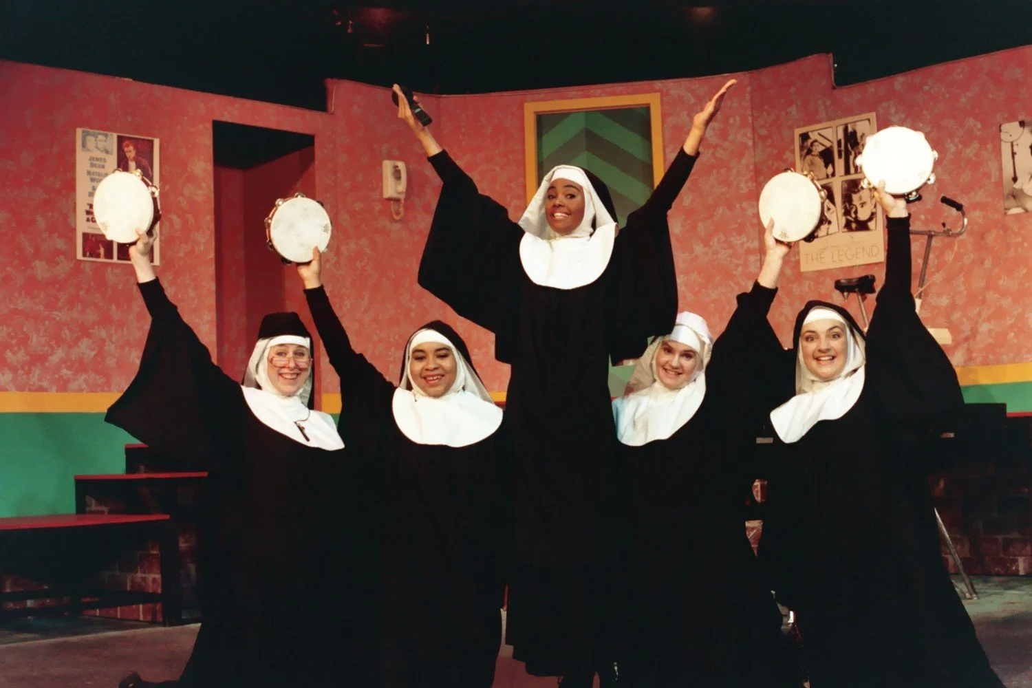 Five people dressed as nuns performing on stage, with three holding tambourines and one raising arms. The background shows a colorful set with posters and musical instruments.