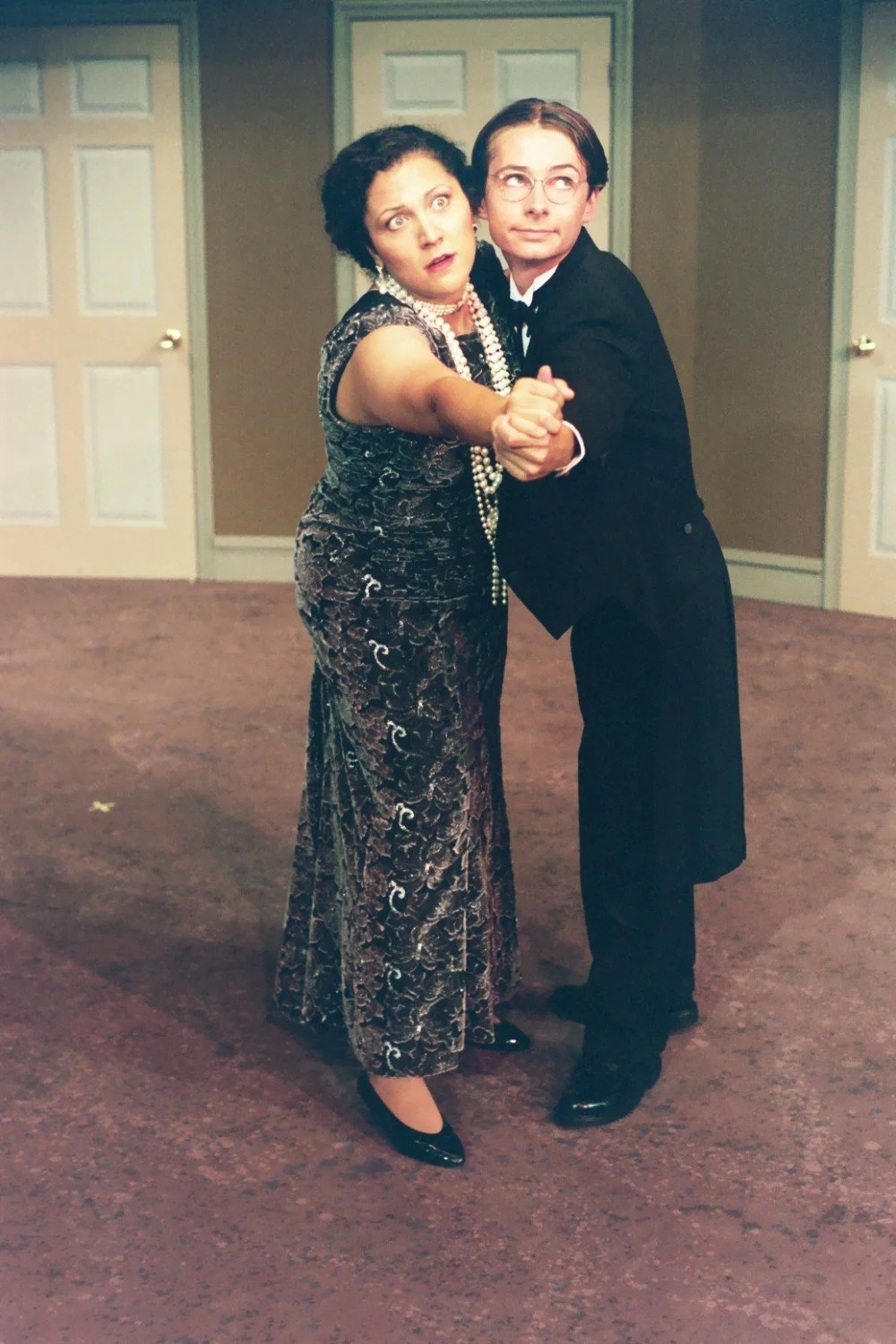 A man and woman dancing in a room with several white doors. The woman is wearing a patterned dress and pearl necklace, while the man is in a tuxedo. Both have surprised expressions.