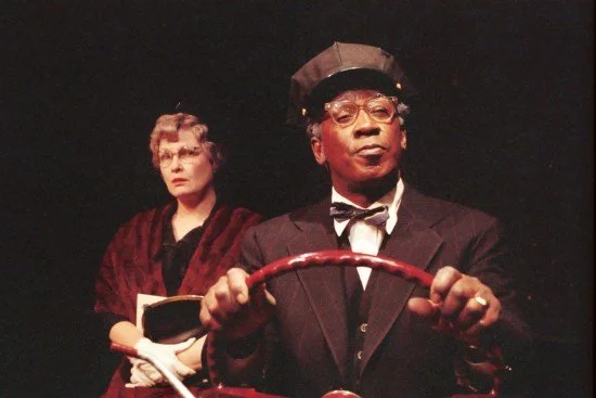 Two actors in a theatrical scene; one is dressed as a chauffeur with a steering wheel, and the other as a passenger, wearing glasses and holding a notebook.