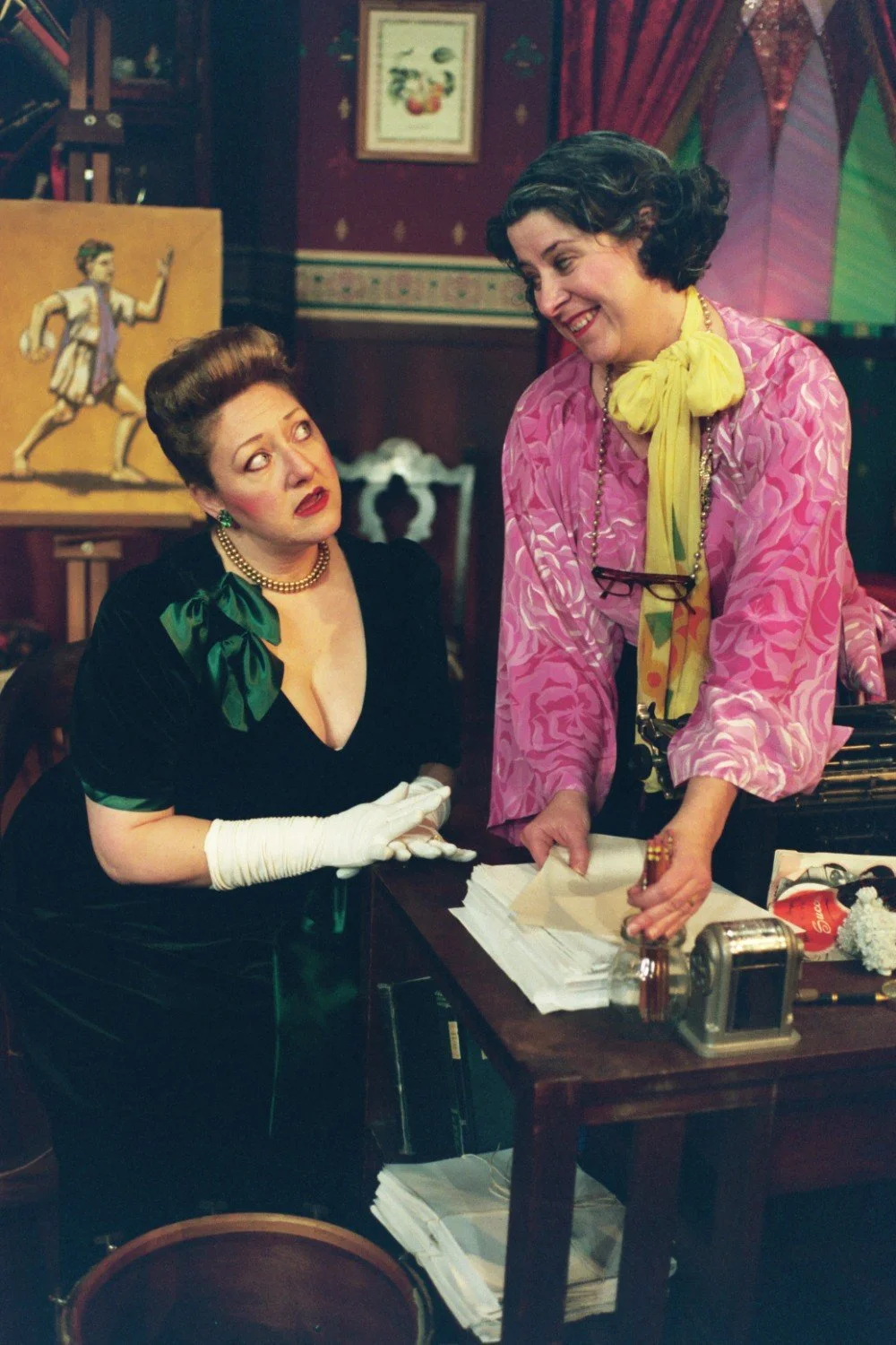 Two women dressed in vintage clothing stand in a room with ornate decor; one sits in a green dress while the other wears a pink blouse.