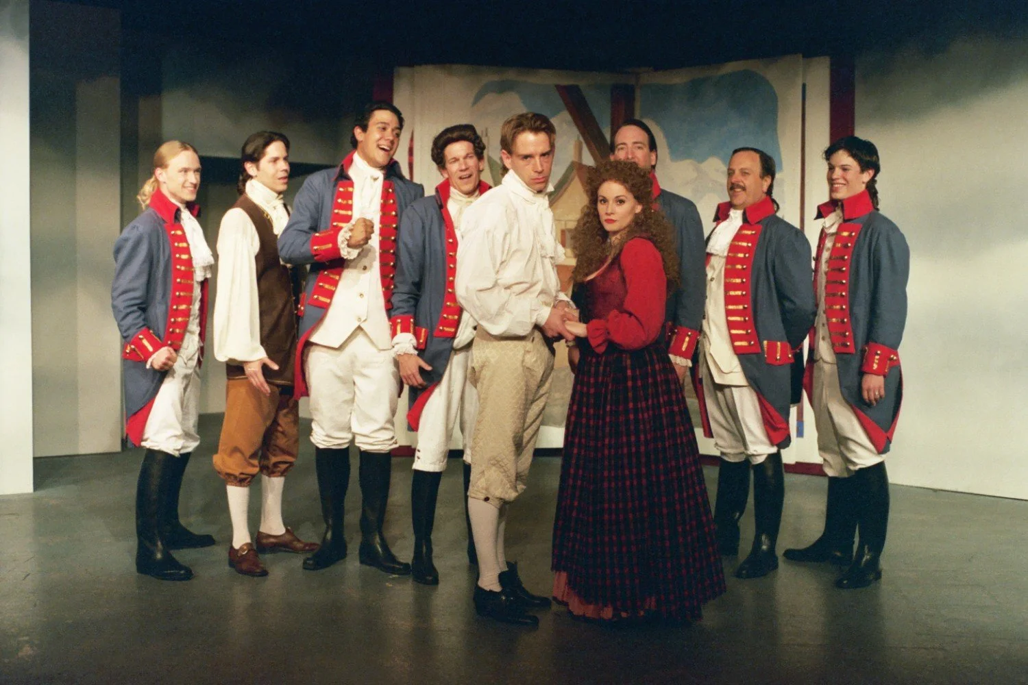 A theatrical performance with actors in period costumes, including men in blue and red military-style uniforms and a woman in a red blouse and long plaid skirt, all standing on stage.
