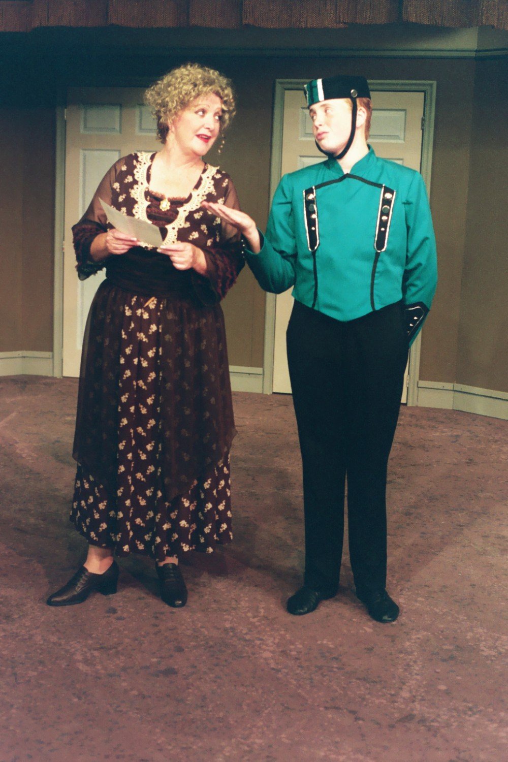 Two actors in costume on stage during a theatrical performance. One is dressed in a vintage dress holding a paper, and the other is in a teal uniform with a cap.
