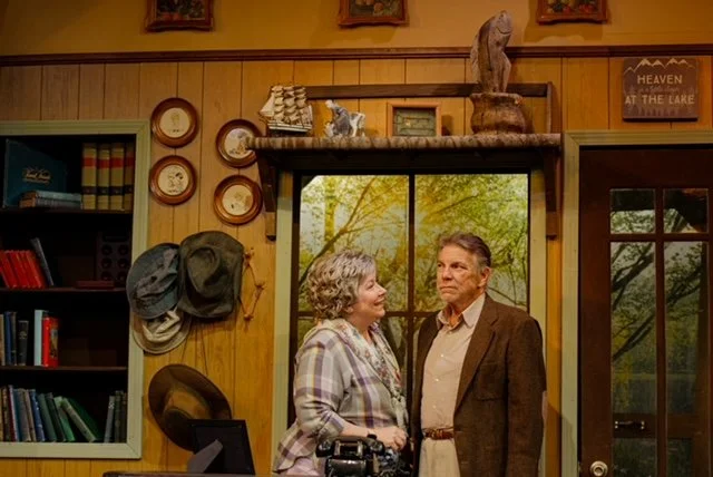 Two people in a cozy room with wood paneling, surrounded by hats on the wall, bookshelves, and nature-themed decor. A window shows a view of trees, creating a rustic atmosphere.
