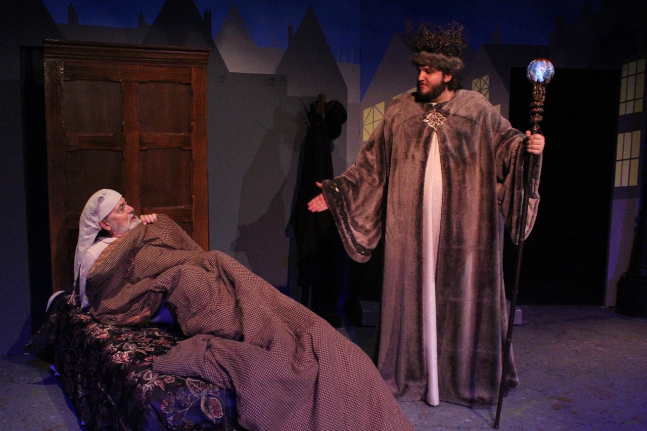 Stage scene from "A Christmas Carol" with Scrooge in bed and a spirit in a robe holding a staff.