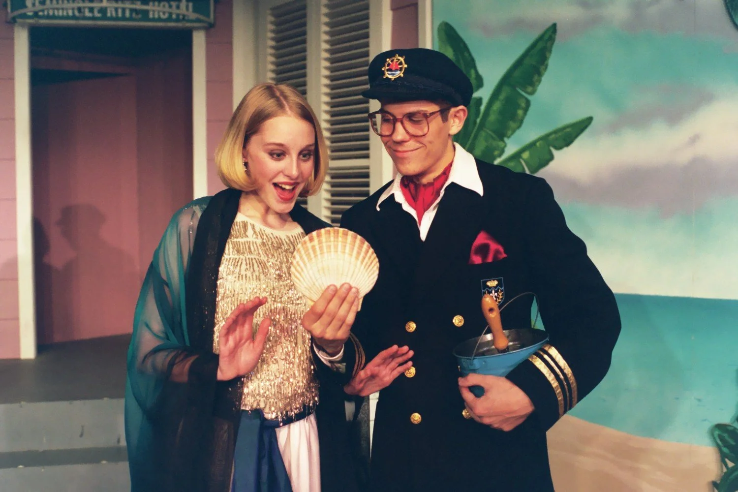 Two people in costume, one holding a seashell, standing in a tropical-themed setting.