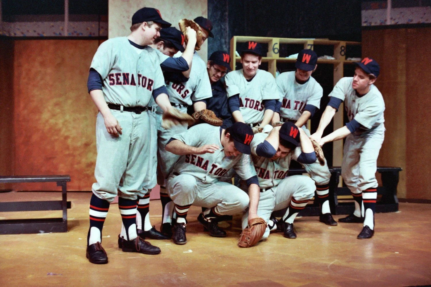 Group of men in vintage baseball uniforms labeled 'Senators' playing around in a locker room setting